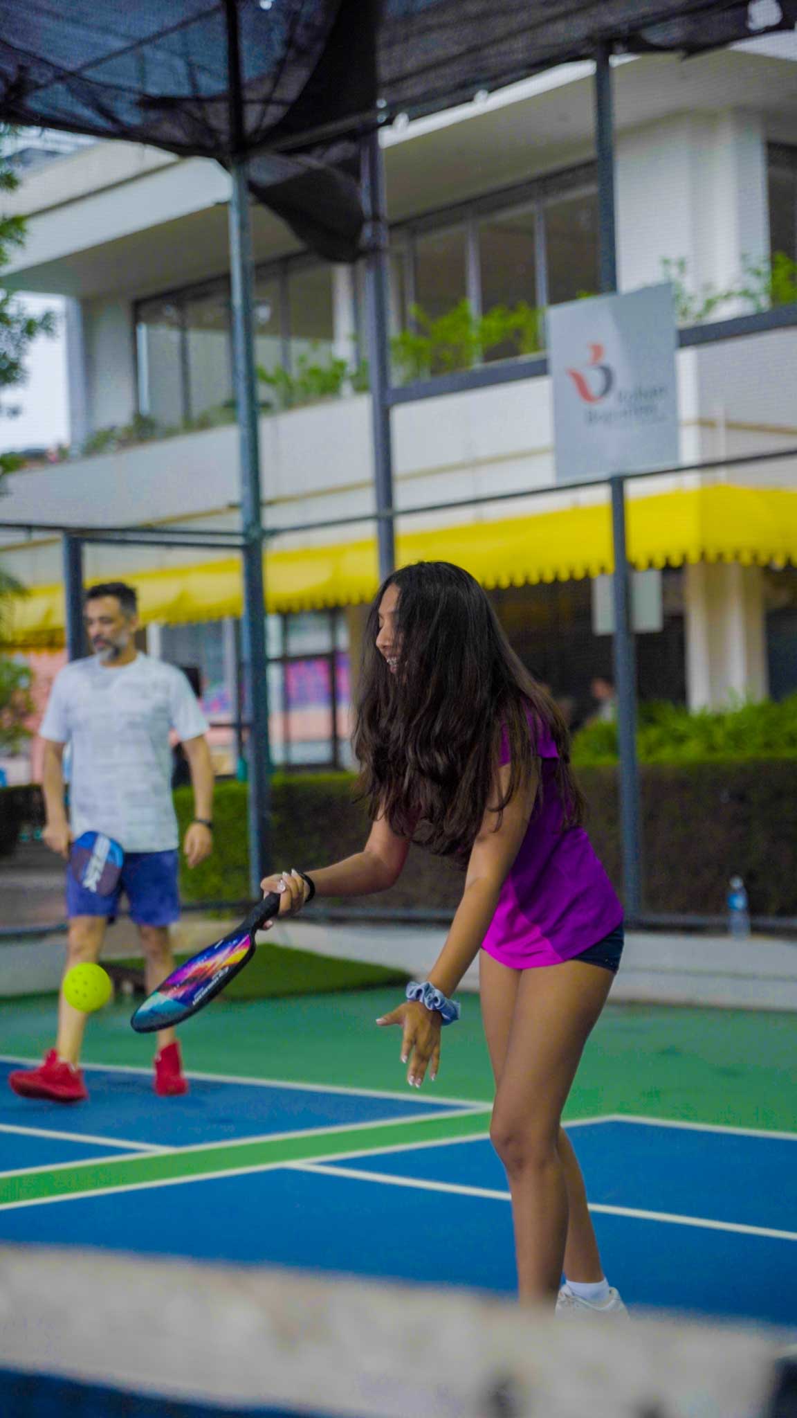 Girl preparing to hit the pickleball with a paddle on the court, man standing behind.
